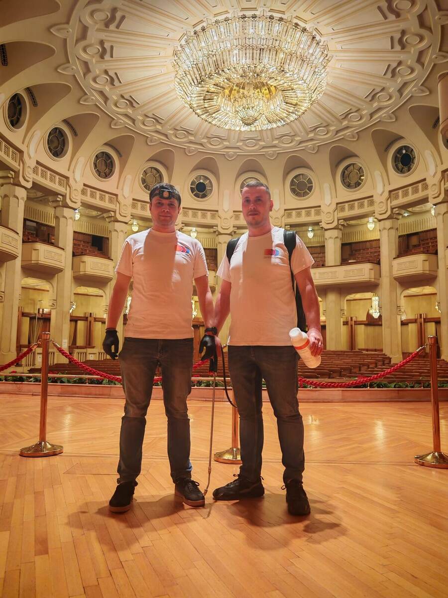 Doi operatori Compania DDD cu pulverizator cu lance și rezervor, într-o sală de spectacole cu candelabru și rânduri de scaune